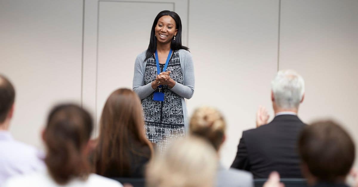 Woman standing at front of room speaking Woman standing at front of room speaking