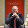 phot0 of Tim Brown sitting on his brick front porch, Welcome sign on a red door behind him
